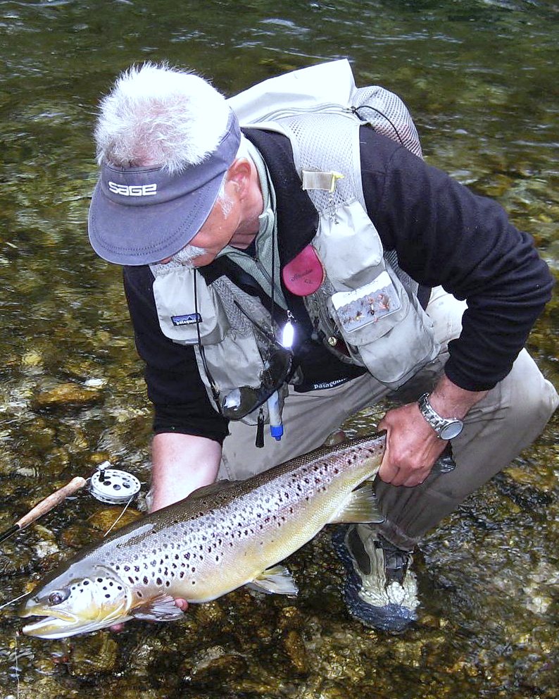 Gerhard Laible in Fischerweste und Schirmkappe kniet in der Deutschen Traun und hält eine große Bachforelle über das Wasser